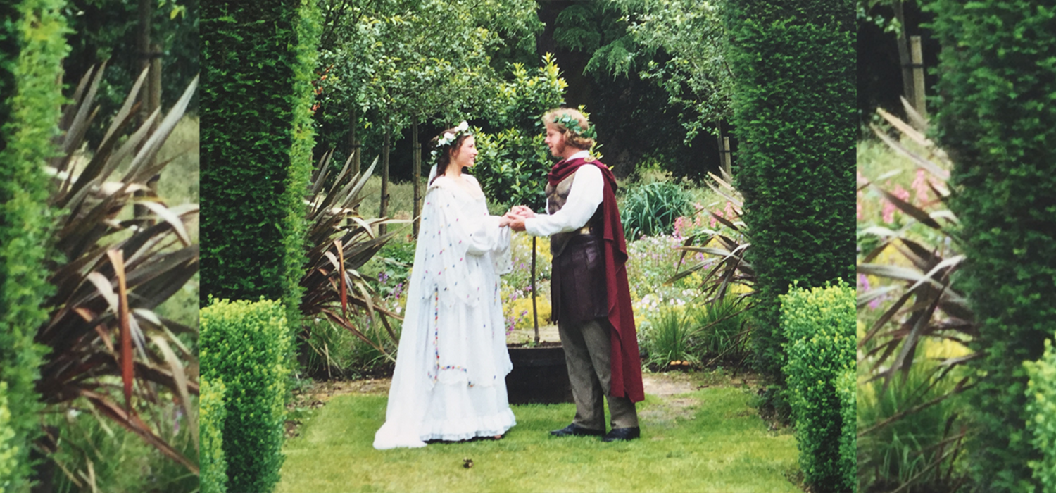 Alumnus Jonathan Love and an actress stand in front of a wooded area wearing Shakespearean garb. They are holding hands and both have flower crowns.