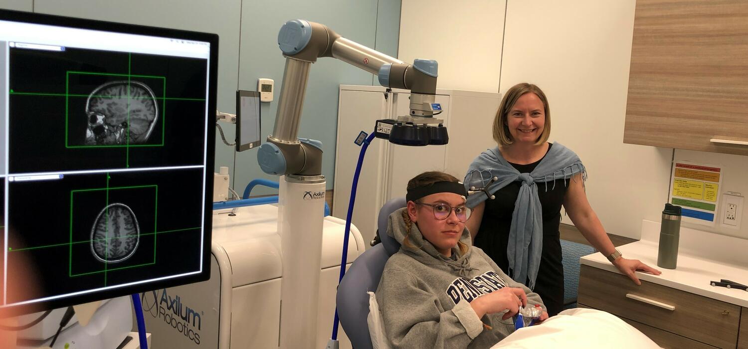 A person sits in a chair in a doctors office while getting a brainscan and another person stands next to them