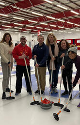 Physicians at the IOC diploma workshop stand behind a curling rock holding curling brooms.