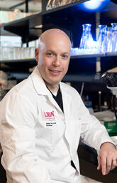 A man in a lab coat sits at a table with a microscope and smiles at the camera